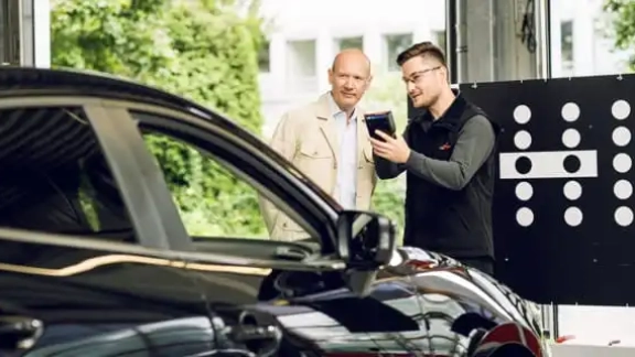 Monteur erklärt Kunde die Kalibrierung eines Fahrzeugs. Monteur hält Gerät in der Hand und zeigt auf das Auto